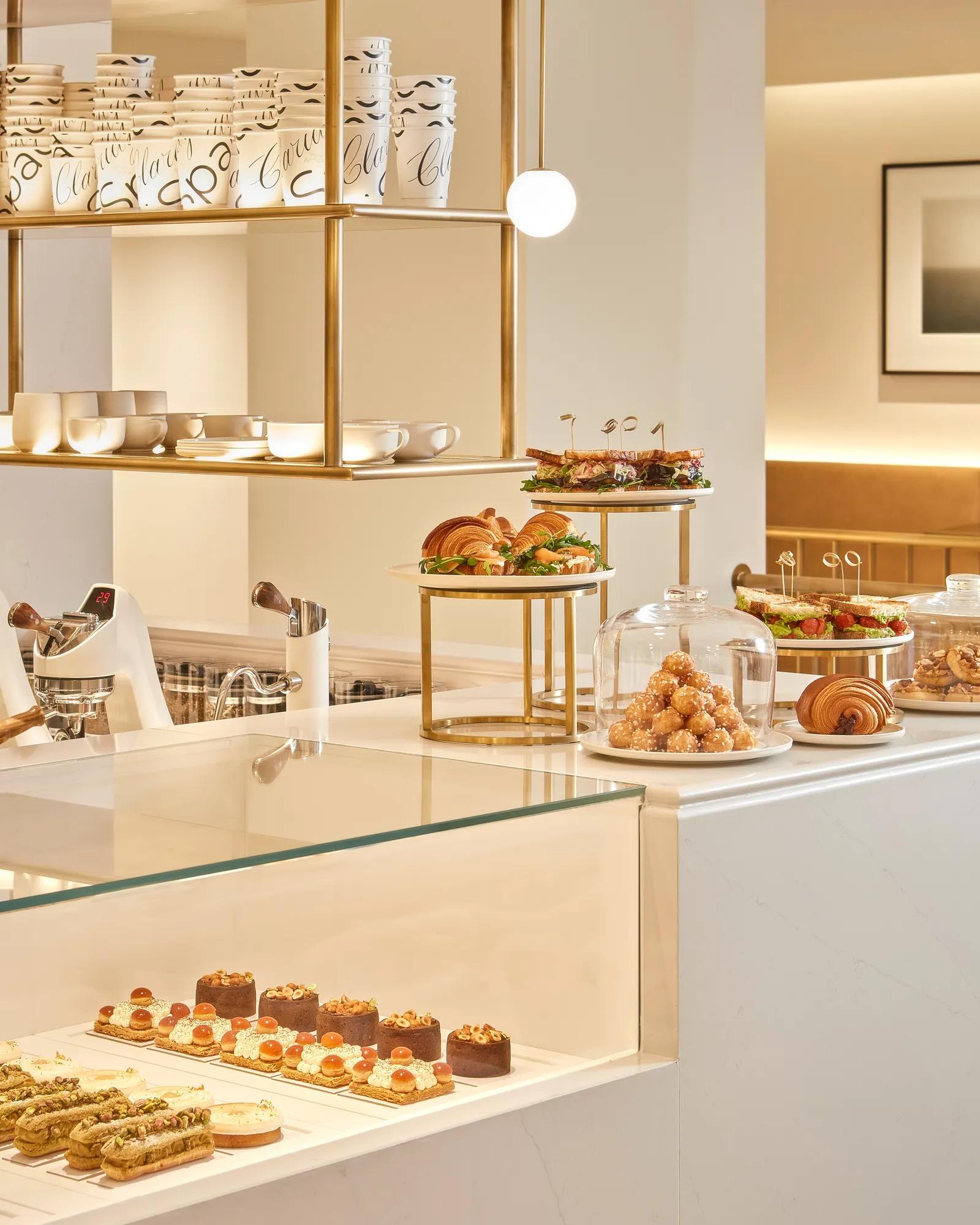 Bright café counter displaying an assortment of elegant pastries, sandwiches, and croissants under glass domes and on tiered stands, with gold shelving and white marble surfaces in a modern setting.