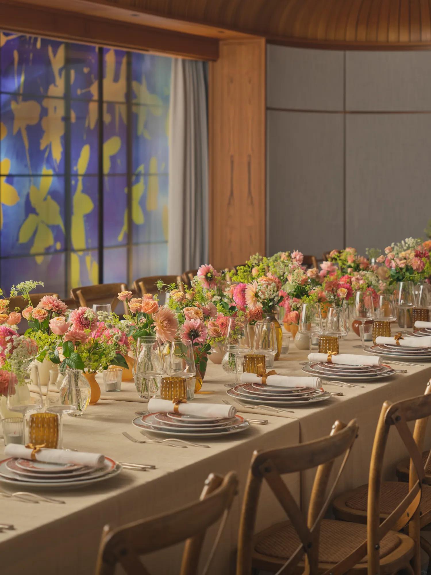 Long dining table set for an event, decorated with pink and peach flowers and glowing amber glassware