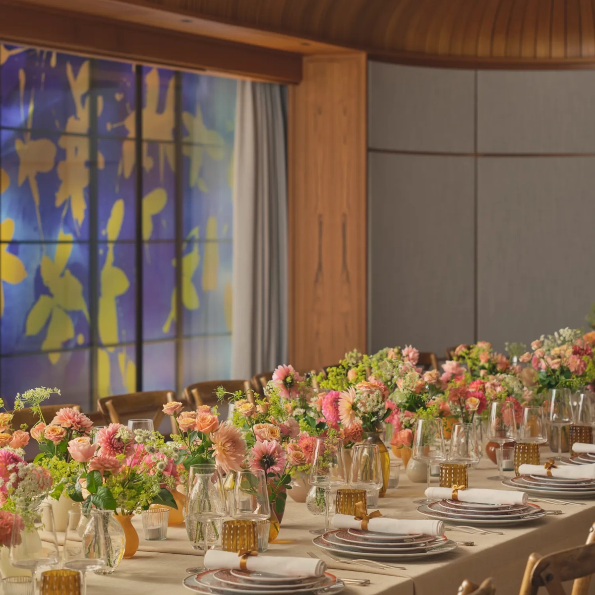 Long dining table set for an event, decorated with pink and peach flowers and glowing amber glassware