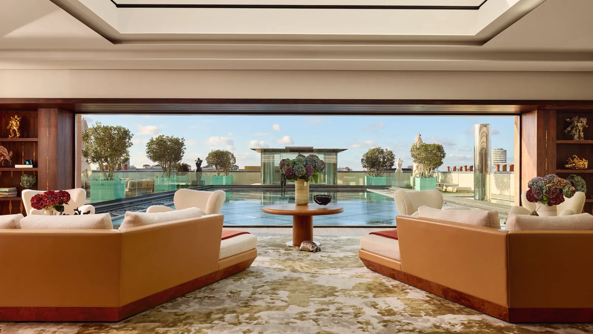 Spacious penthouse living room at Claridge’s with large sofas facing a glass wall opening onto a rooftop terrace and reflective pool.