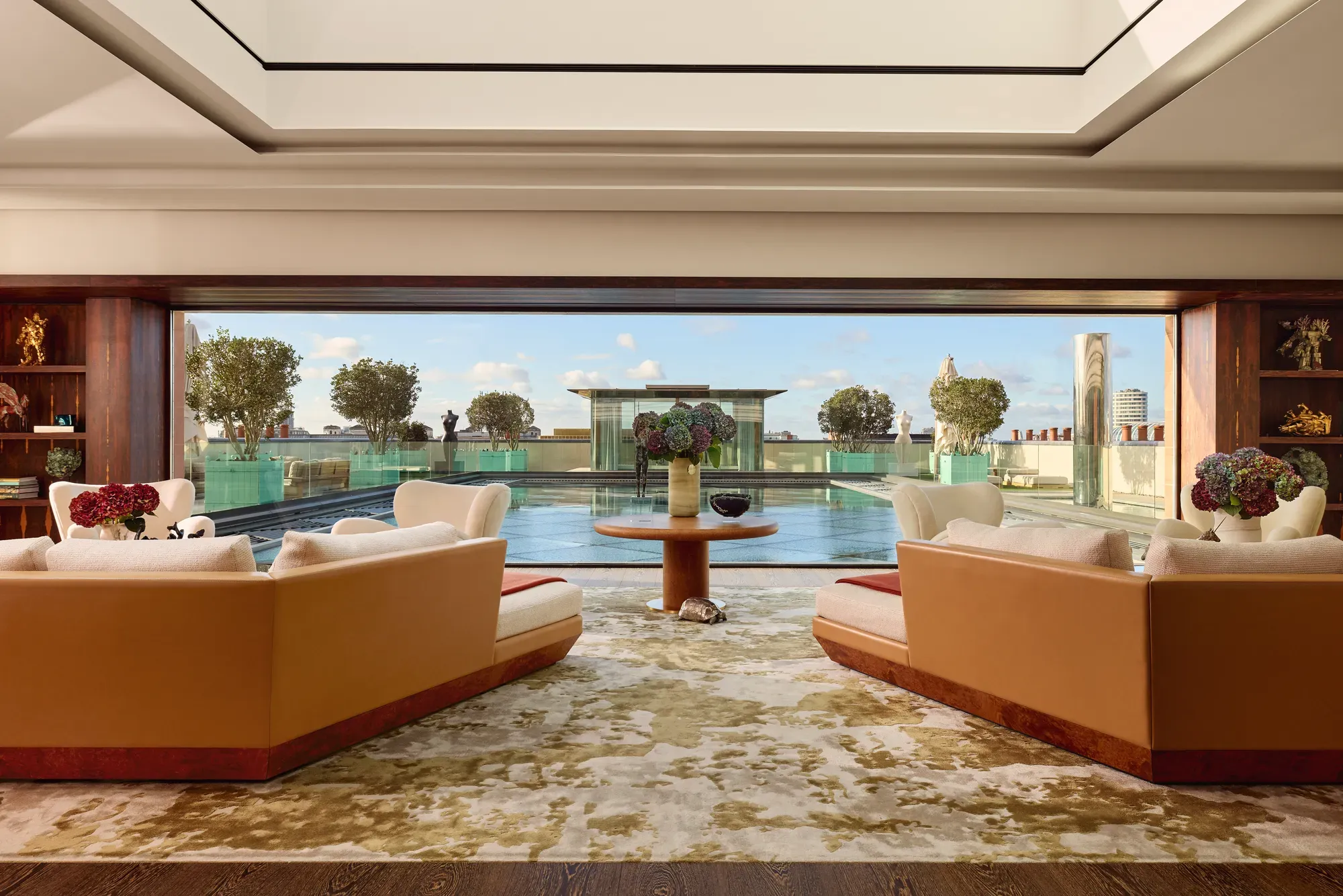 Spacious penthouse living room at Claridge’s with large sofas facing a glass wall opening onto a rooftop terrace and reflective pool.