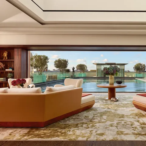 Spacious penthouse living room at Claridge’s with large sofas facing a glass wall opening onto a rooftop terrace and reflective pool.