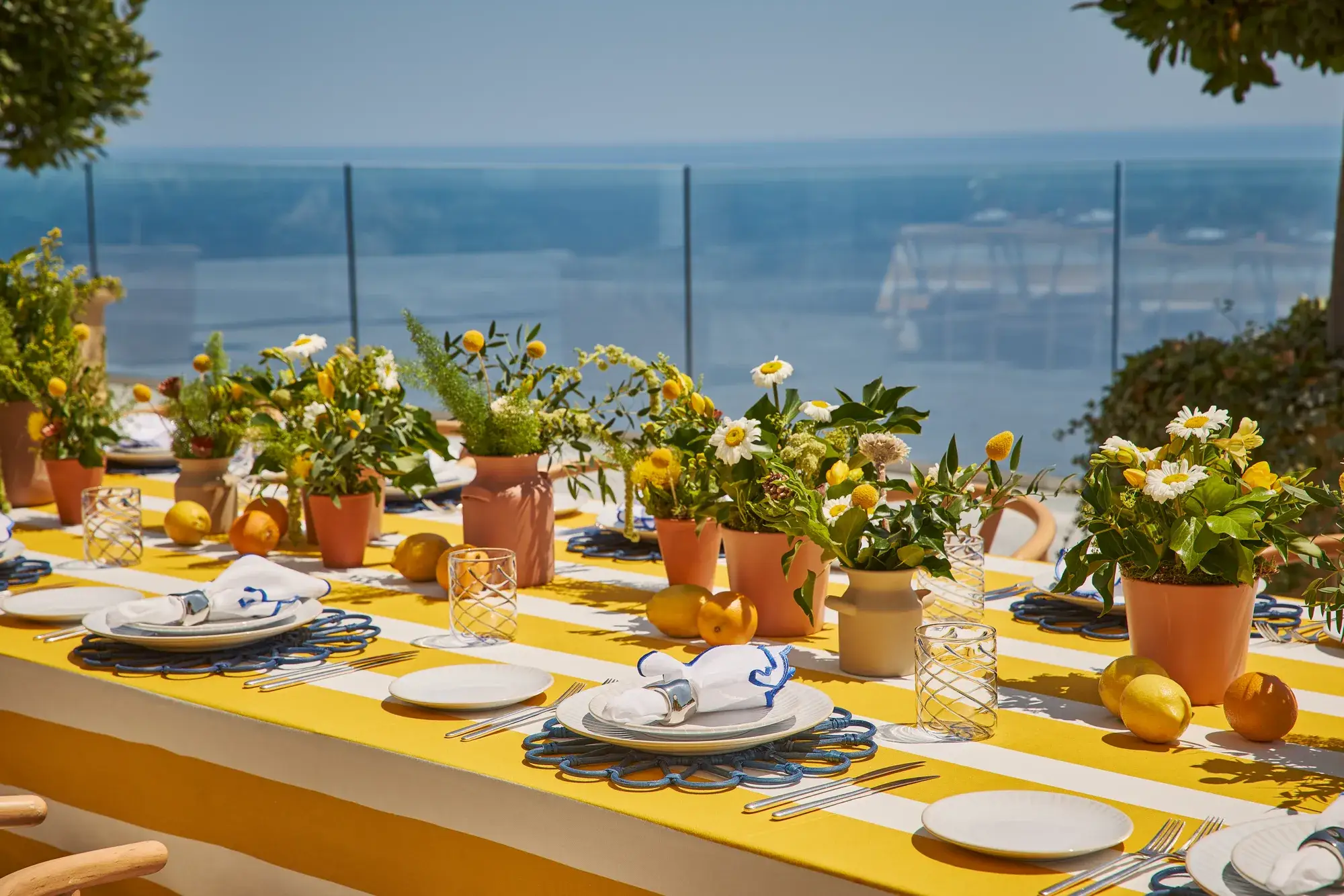 An outdoor dining table with yellow-and-white striped cloth, decorated with potted flowers, lemons, and neatly set plates overlooking the sea.