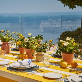 An outdoor dining table with yellow-and-white striped cloth, decorated with potted flowers, lemons, and neatly set plates overlooking the sea.