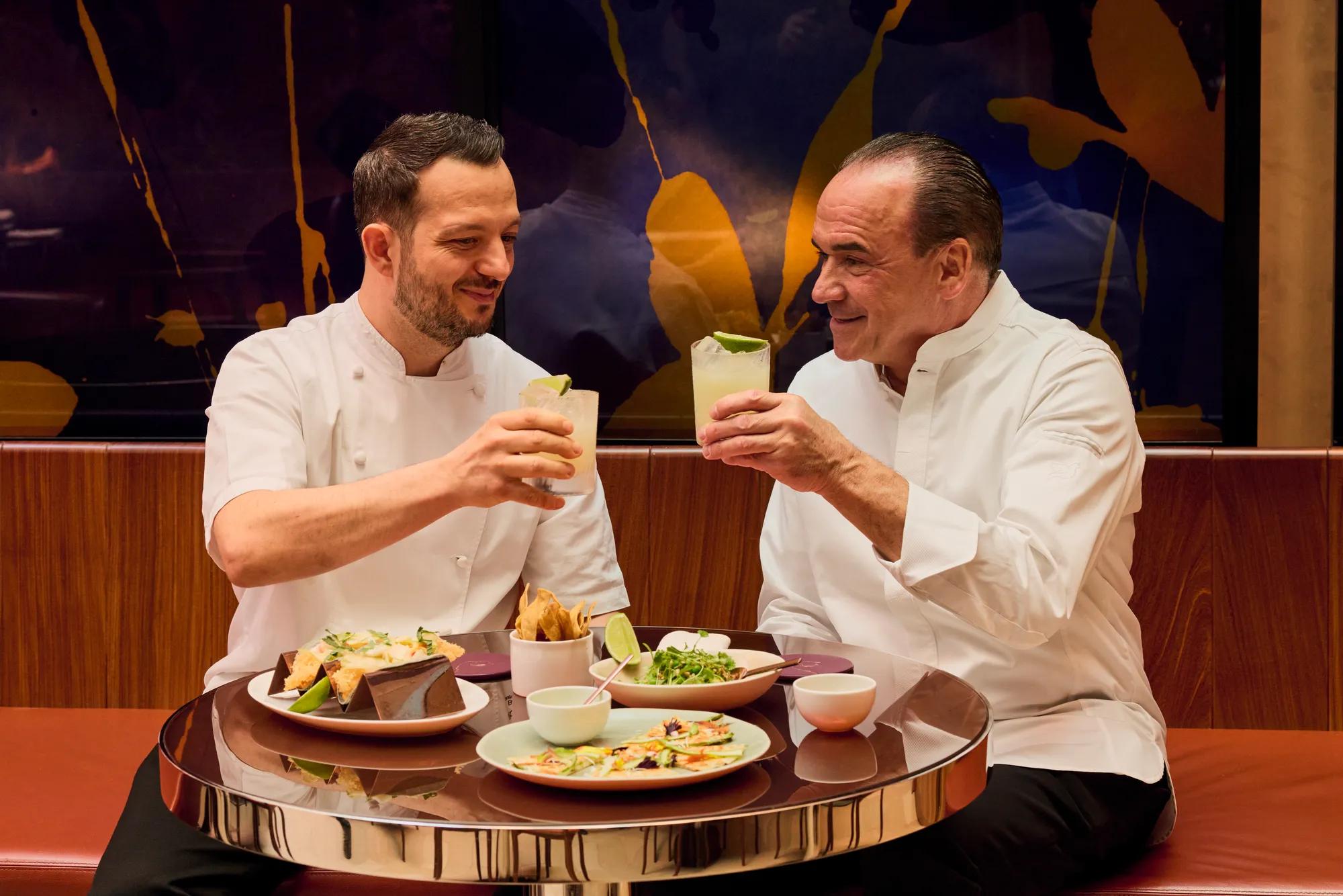 Two chefs raise cocktails over tacos and shared plates at abc kitchens restaurant.
