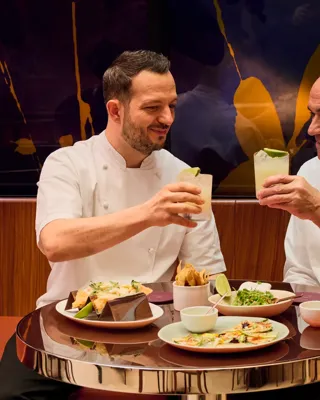 Two chefs raise cocktails over tacos and shared plates at abc kitchens restaurant.