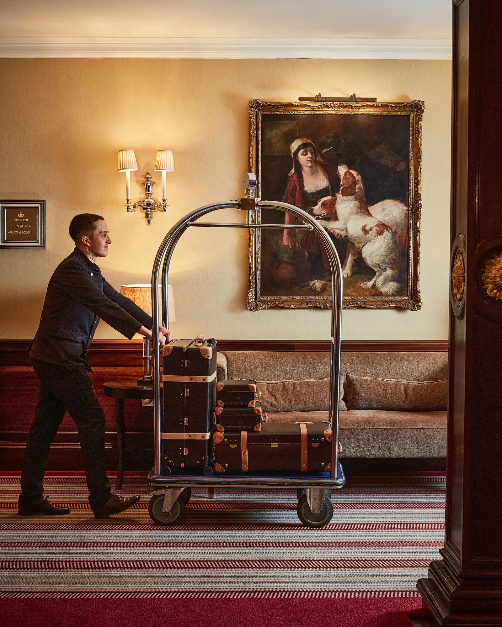A hotel porter pushes a luggage trolley stacked with vintage-style suitcases through an elegant hallway with classic artwork and warm lighting.