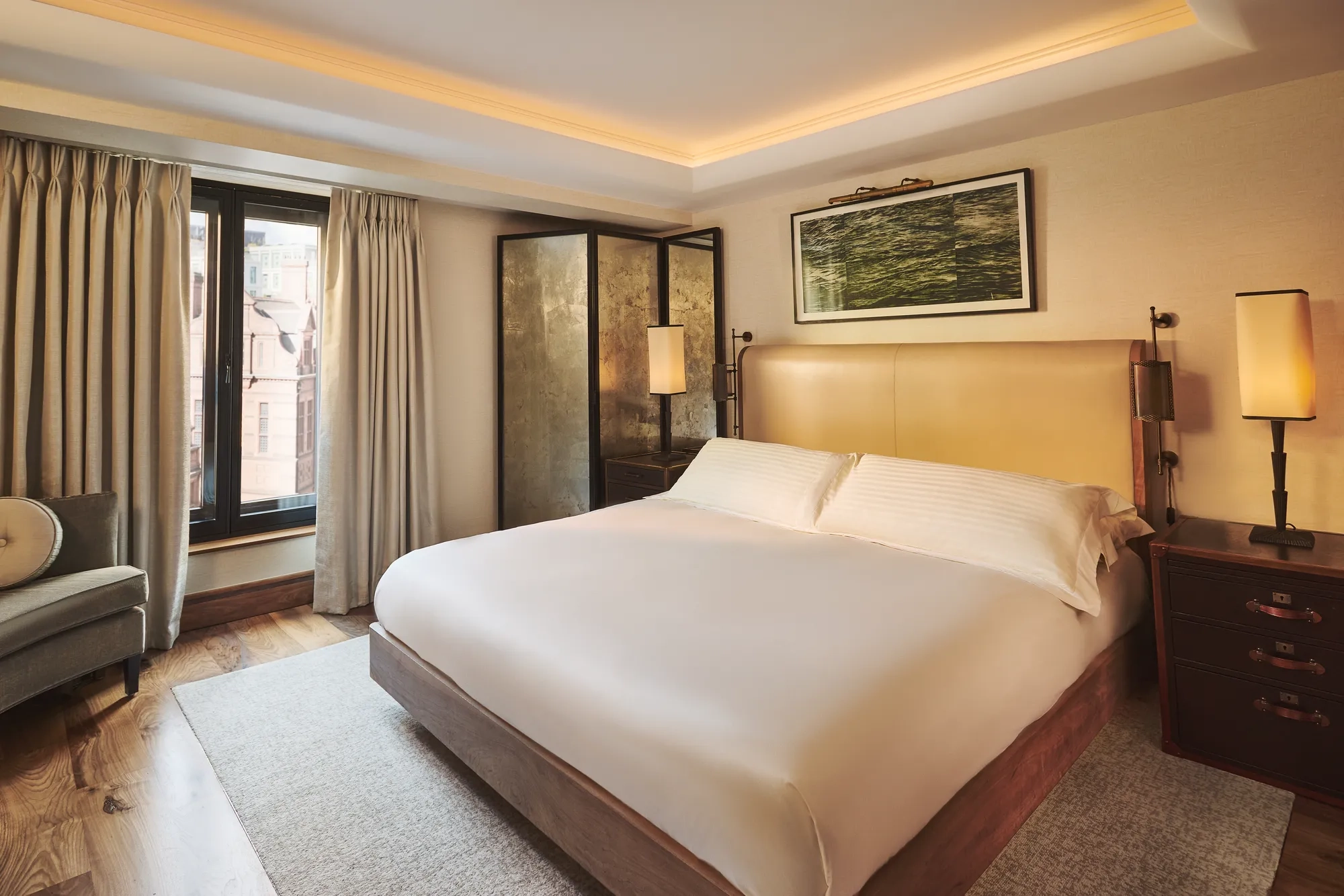 Warm, modern bedroom with a large bed, beige upholstered headboard, bedside lamps, and floor-to-ceiling curtains by tall windows.