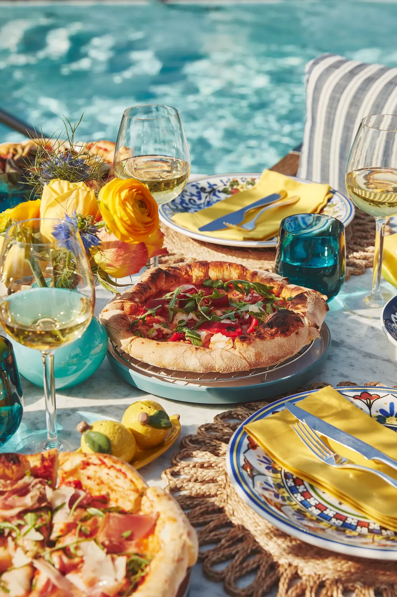 Outdoor table by a pool with pizzas, white wine, yellow napkins, and bright flowers in sunlight.