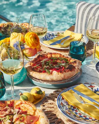 Outdoor table by a pool with pizzas, white wine, yellow napkins, and bright flowers in sunlight.