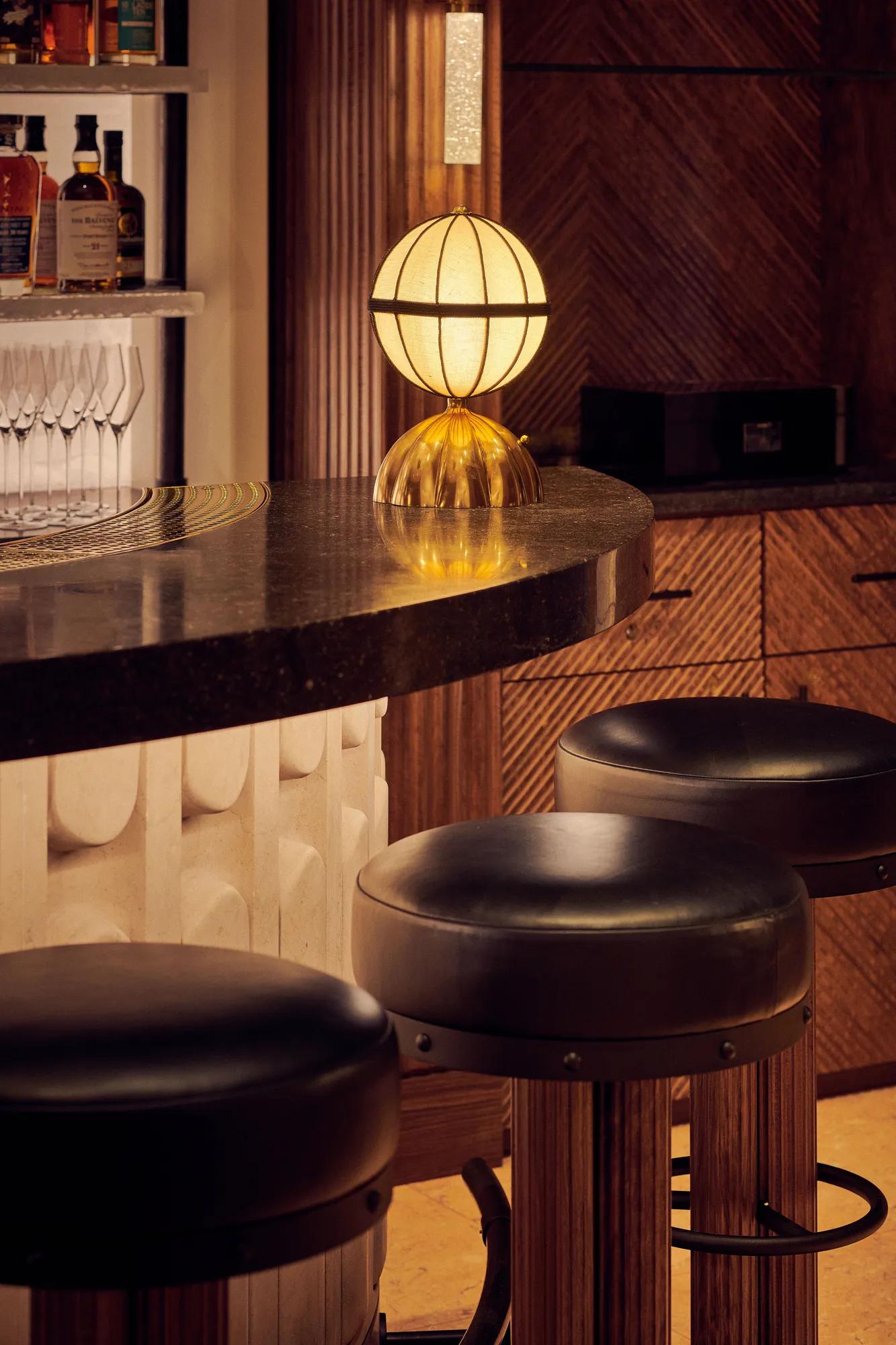 Close-up of bar counter with golden globe lamp and leather bar stools in warm lighting.