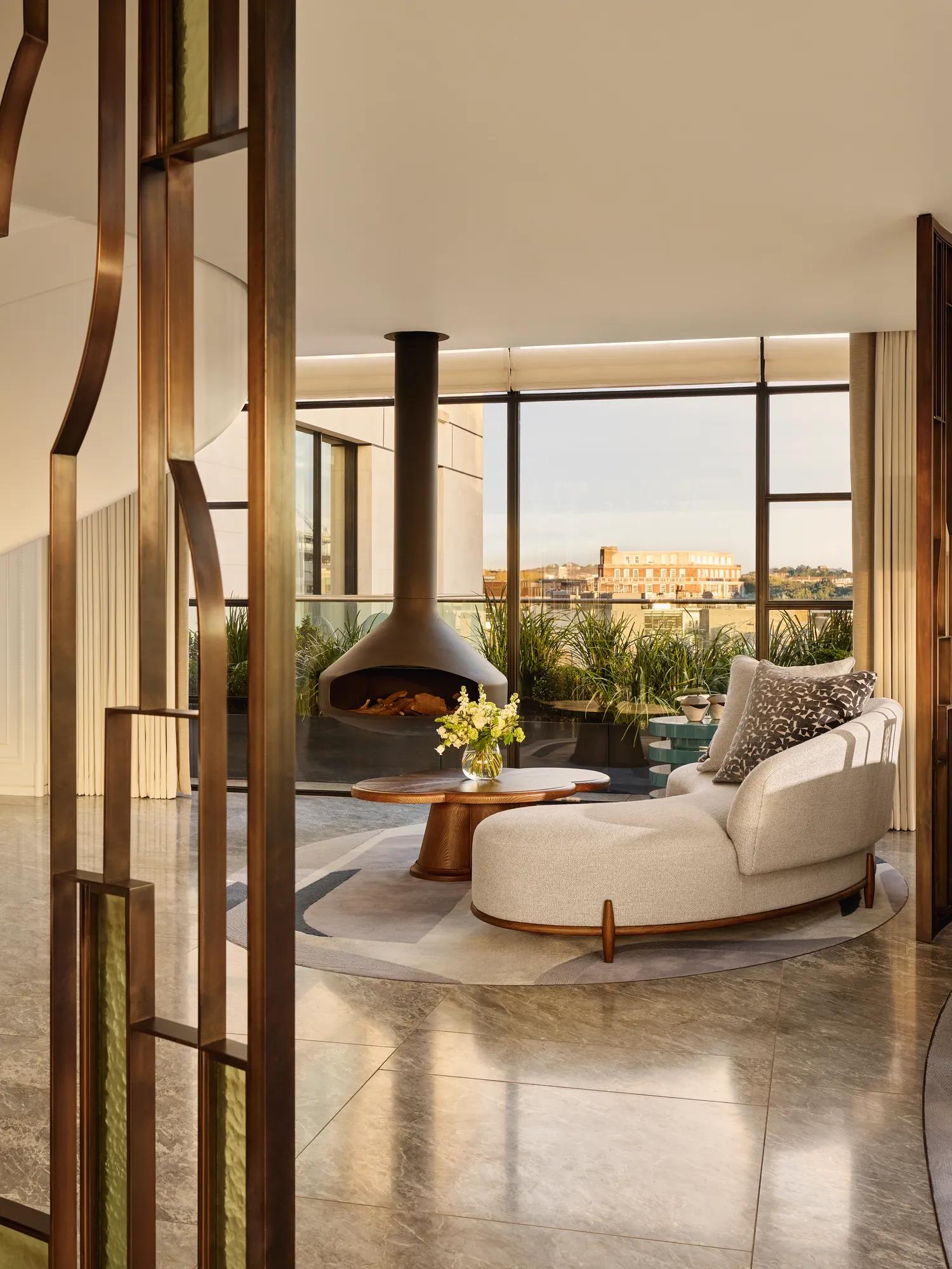 Contemporary living space with a curved cream sofa and round wooden coffee table beside a suspended black fireplace, framed by floor-to-ceiling windows overlooking a city terrace with greenery.