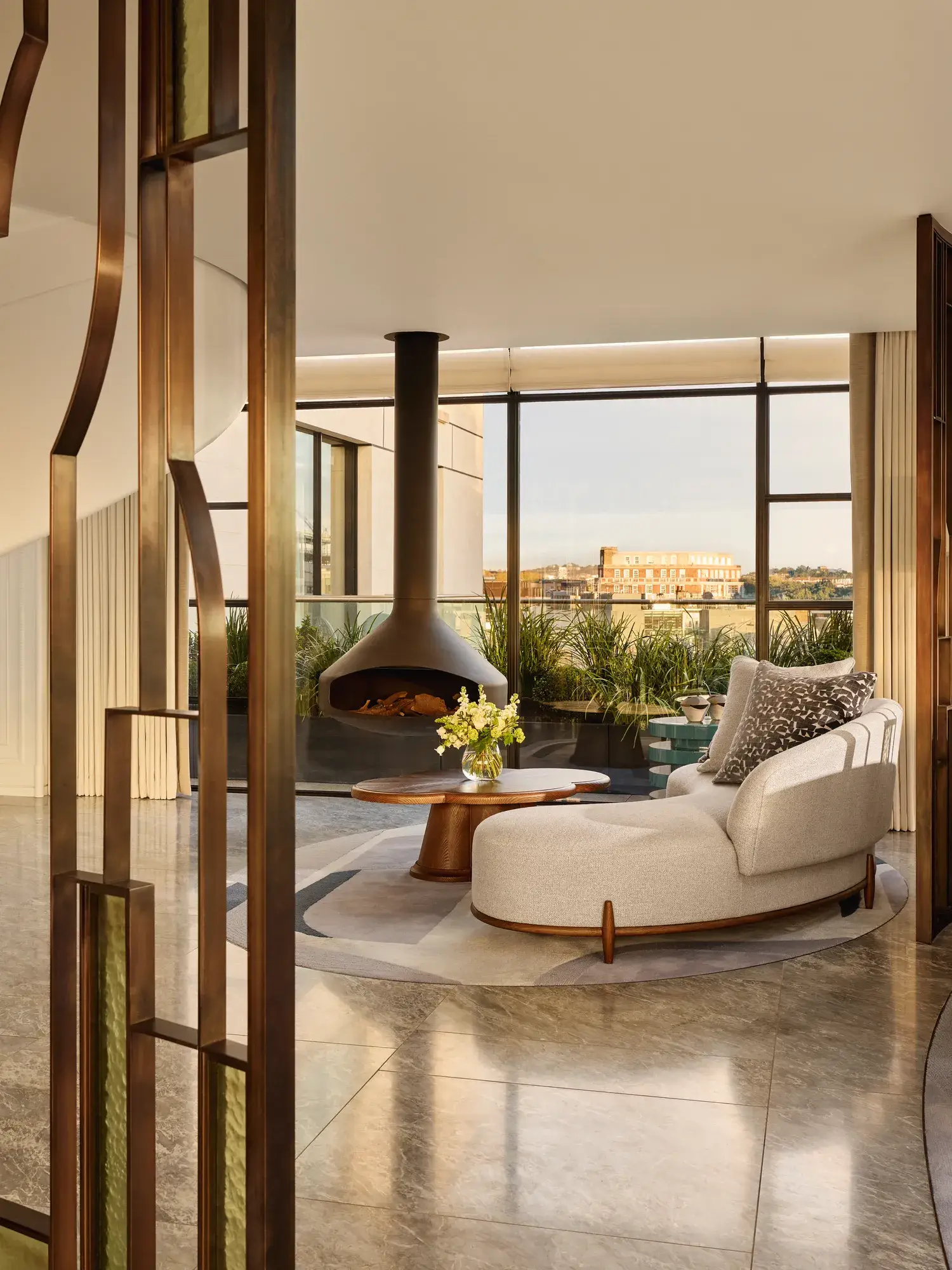 Contemporary living space with a curved cream sofa and round wooden coffee table beside a suspended black fireplace, framed by floor-to-ceiling windows overlooking a city terrace with greenery.