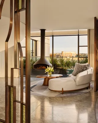 Contemporary living space with a curved cream sofa and round wooden coffee table beside a suspended black fireplace, framed by floor-to-ceiling windows overlooking a city terrace with greenery.