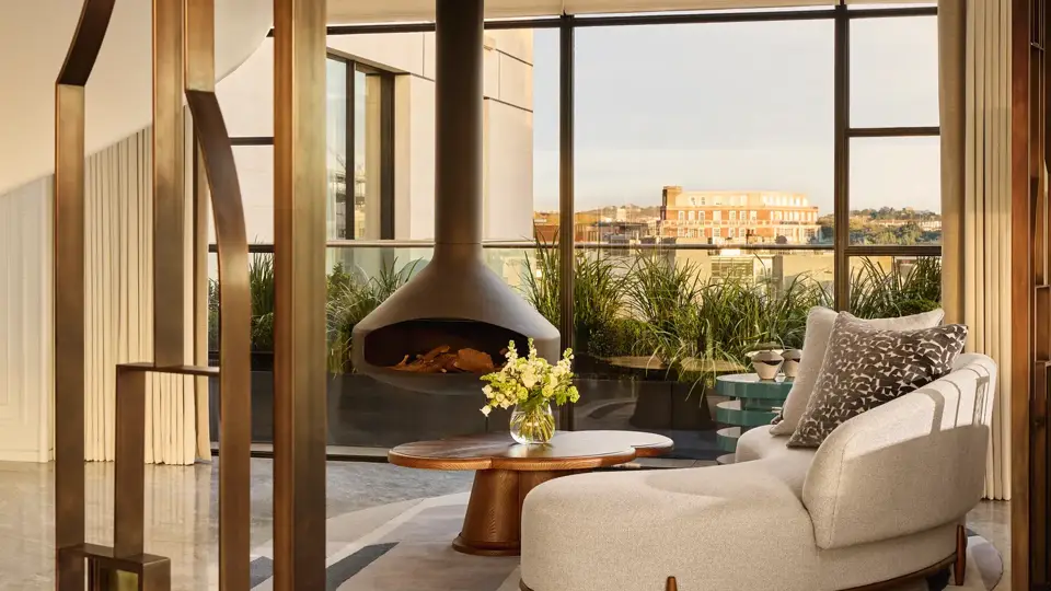 Contemporary living space with a curved cream sofa and round wooden coffee table beside a suspended black fireplace, framed by floor-to-ceiling windows overlooking a city terrace with greenery.