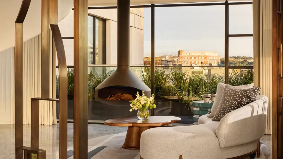 Contemporary living space with a curved cream sofa and round wooden coffee table beside a suspended black fireplace, framed by floor-to-ceiling windows overlooking a city terrace with greenery.