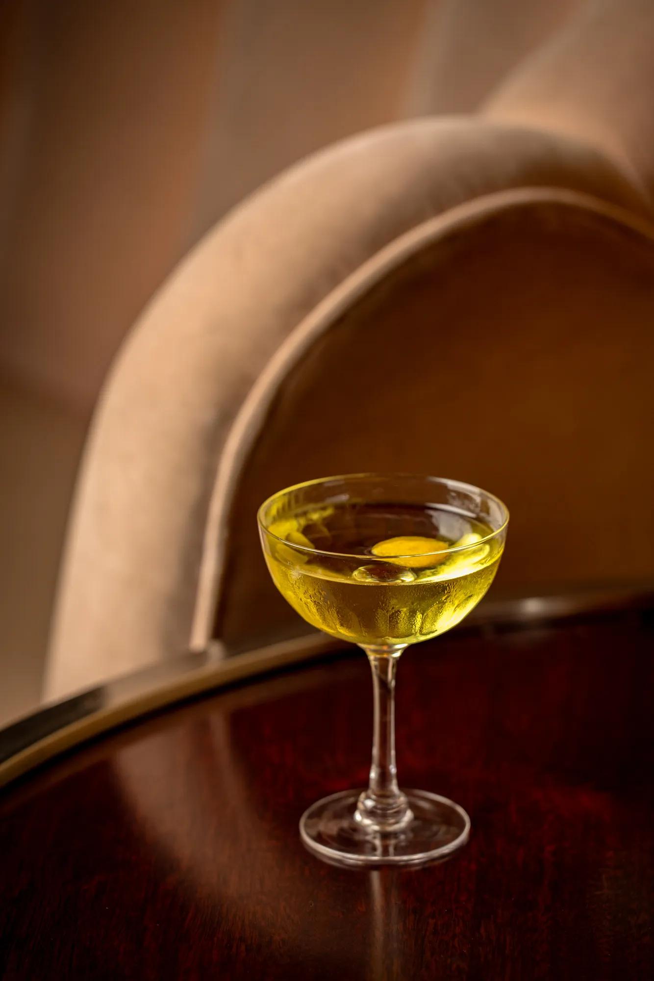 Yellow cocktail in a coupe glass on a polished wooden table, with curved upholstered chair softly blurred in the background.