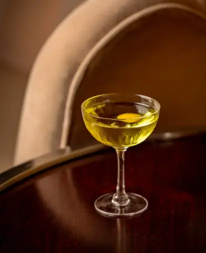 Yellow cocktail in a coupe glass on a polished wooden table, with curved upholstered chair softly blurred in the background.