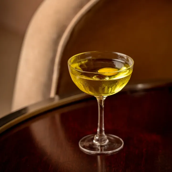 Yellow cocktail in a coupe glass on a polished wooden table, with curved upholstered chair softly blurred in the background.