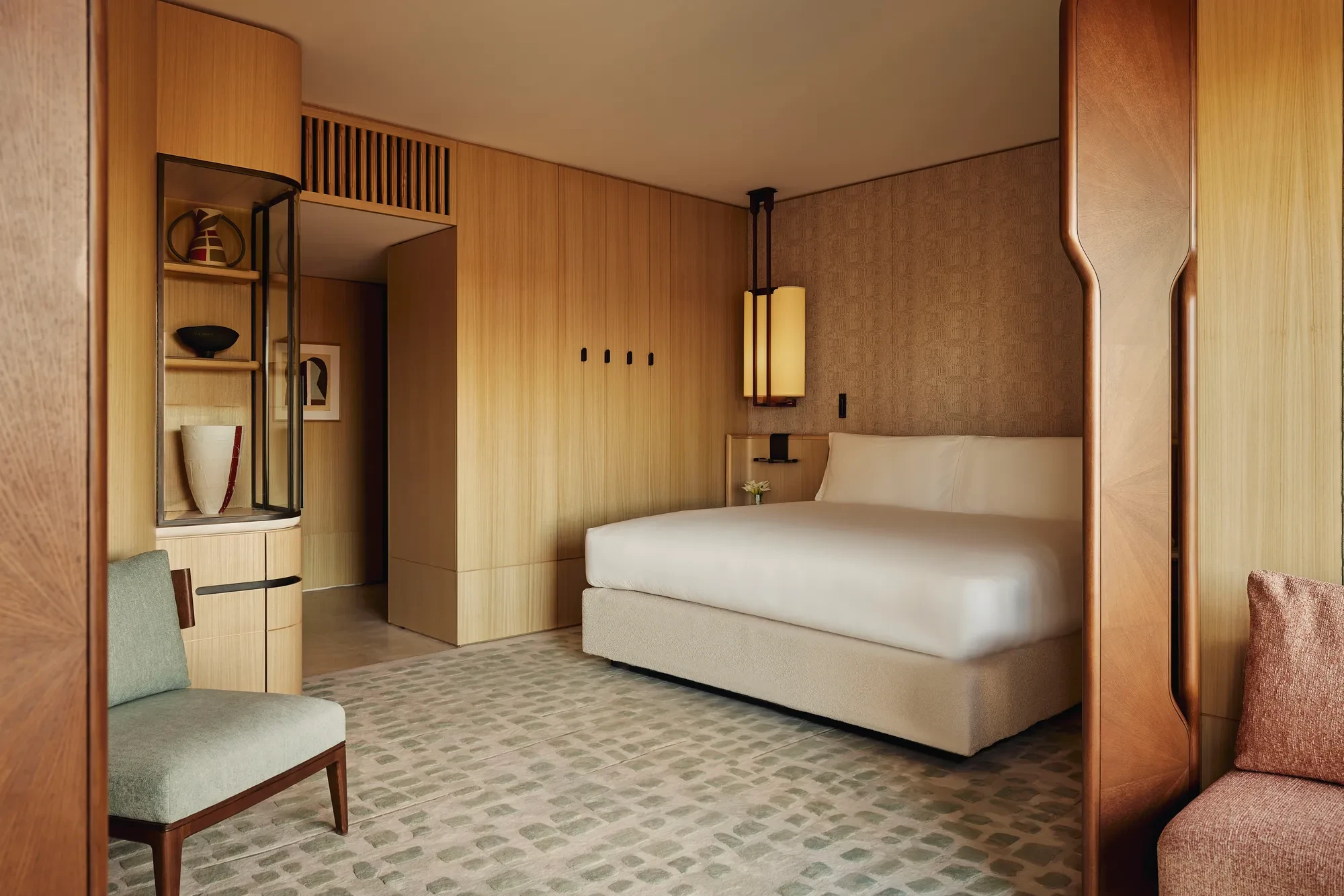 Hotel bedroom with a large bed, soft patterned carpet, light wood panelled walls, and a green upholstered chair in the foreground.