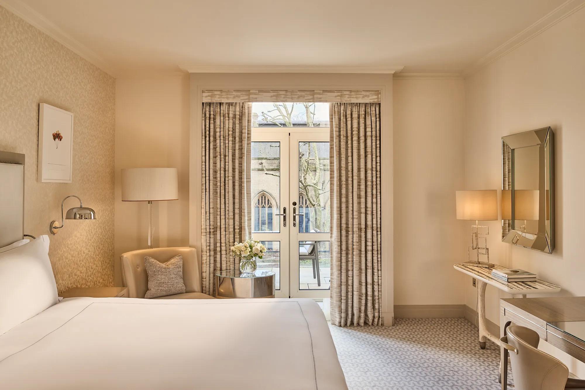 Beige guest room with bed, armchair and desk facing French doors with curtains opening to a small balcony and city view.