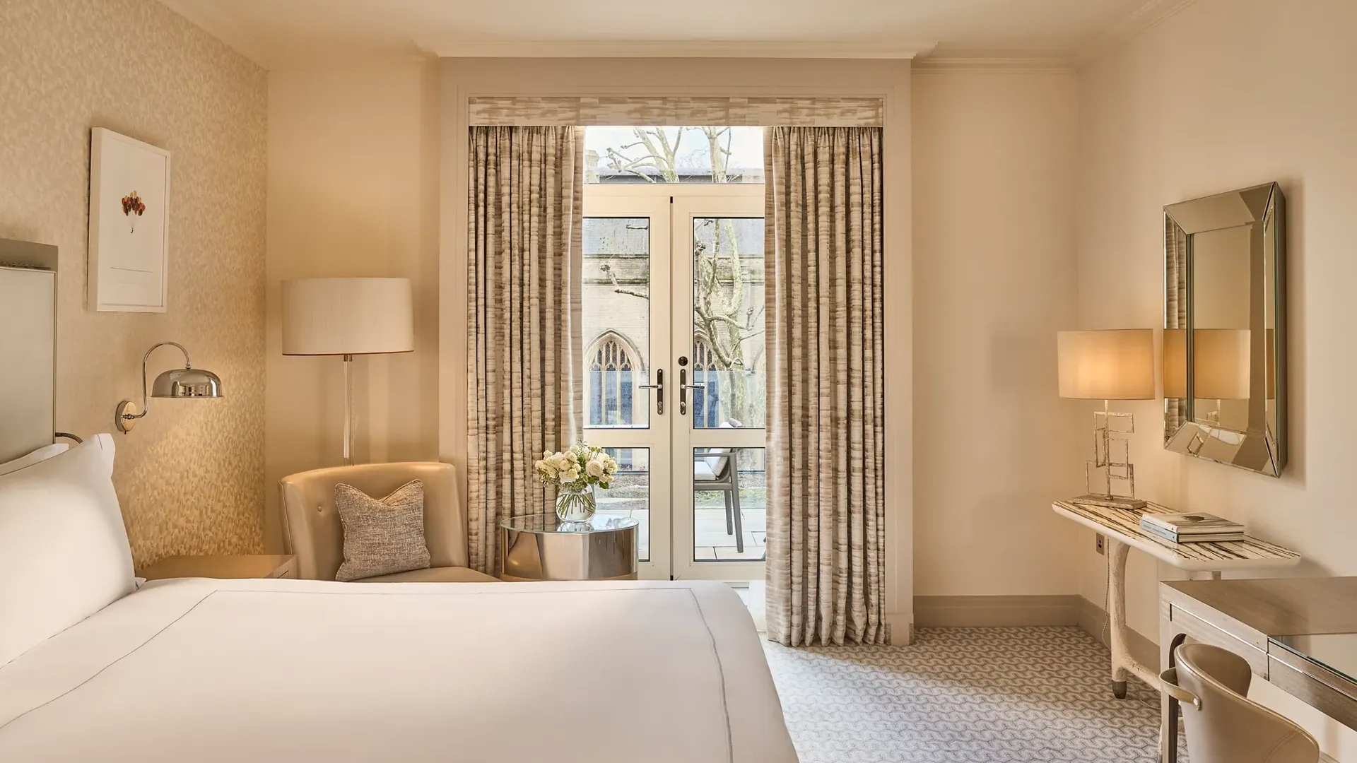 Beige guest room with bed, armchair and desk facing French doors with curtains opening to a small balcony and city view.