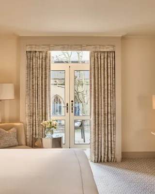 Beige guest room with bed, armchair and desk facing French doors with curtains opening to a small balcony and city view.