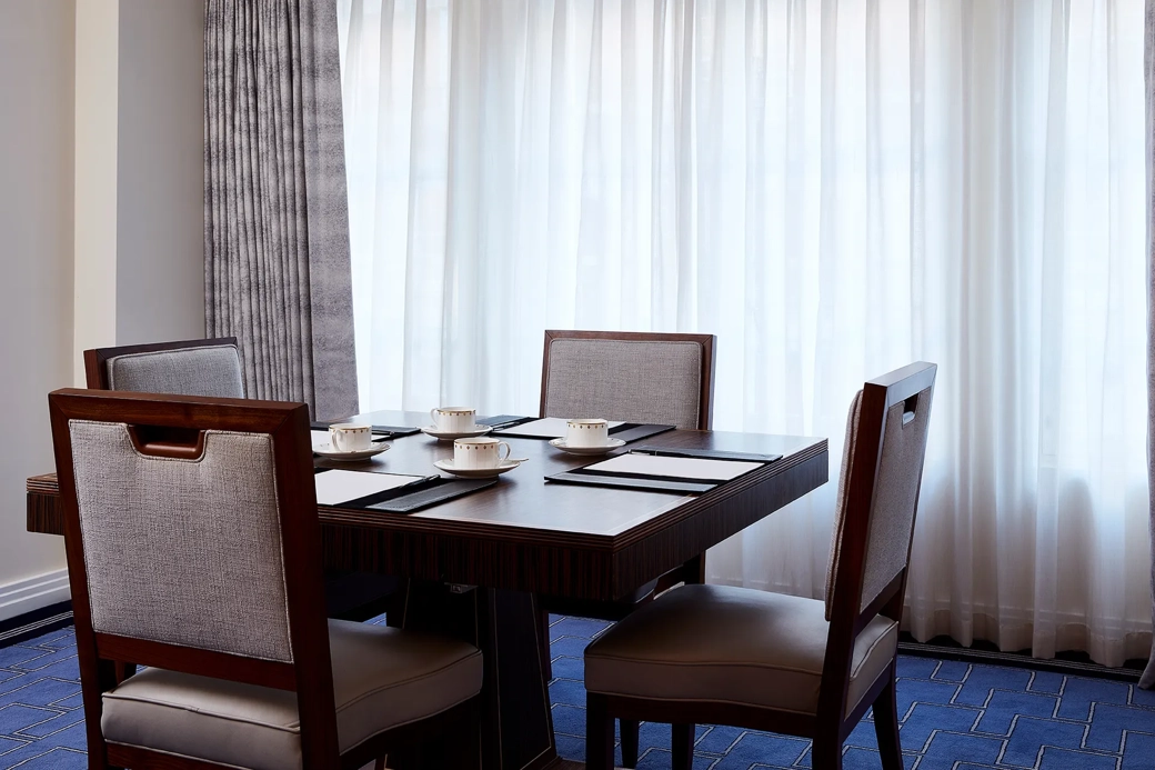 A small meeting setup in the Clarence Room at Claridge’s with four upholstered chairs around a dark wooden table, set with notepads and fine china cups beside tall curtained windows.