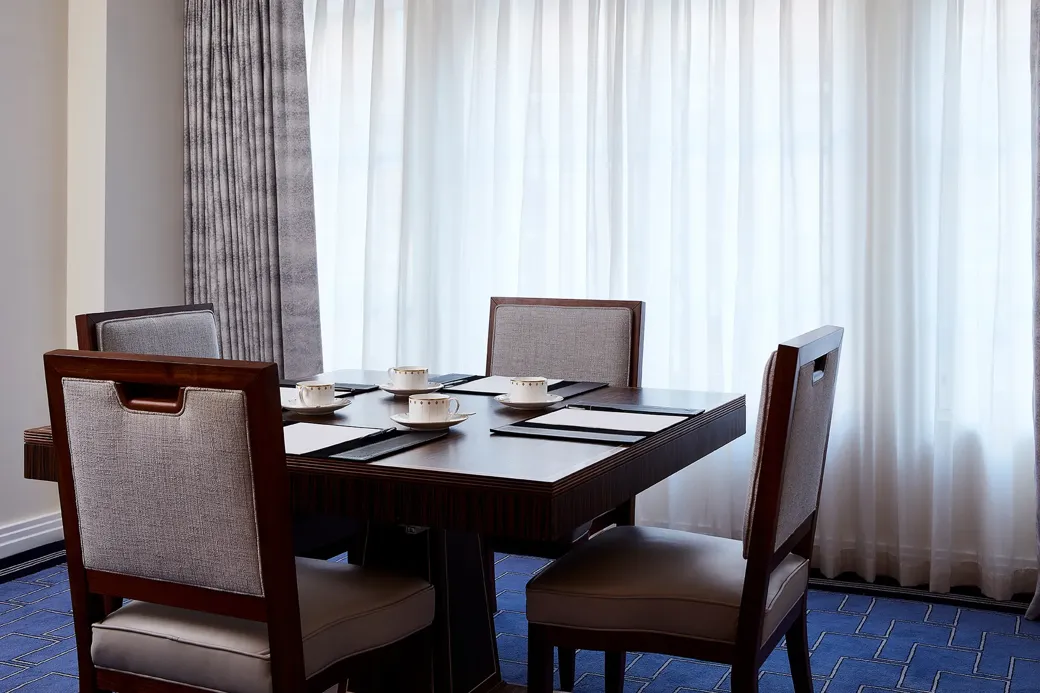 A small meeting setup in the Clarence Room at Claridge’s with four upholstered chairs around a dark wooden table, set with notepads and fine china cups beside tall curtained windows.