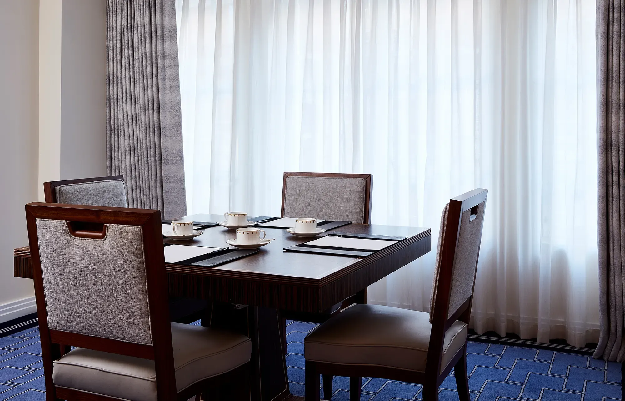 A small meeting setup in the Clarence Room at Claridge’s with four upholstered chairs around a dark wooden table, set with notepads and fine china cups beside tall curtained windows.