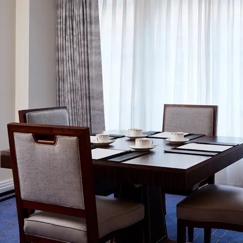 A small meeting setup in the Clarence Room at Claridge’s with four upholstered chairs around a dark wooden table, set with notepads and fine china cups beside tall curtained windows.