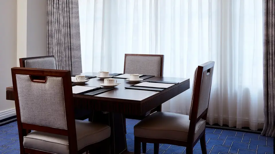 A small meeting setup in the Clarence Room at Claridge’s with four upholstered chairs around a dark wooden table, set with notepads and fine china cups beside tall curtained windows.