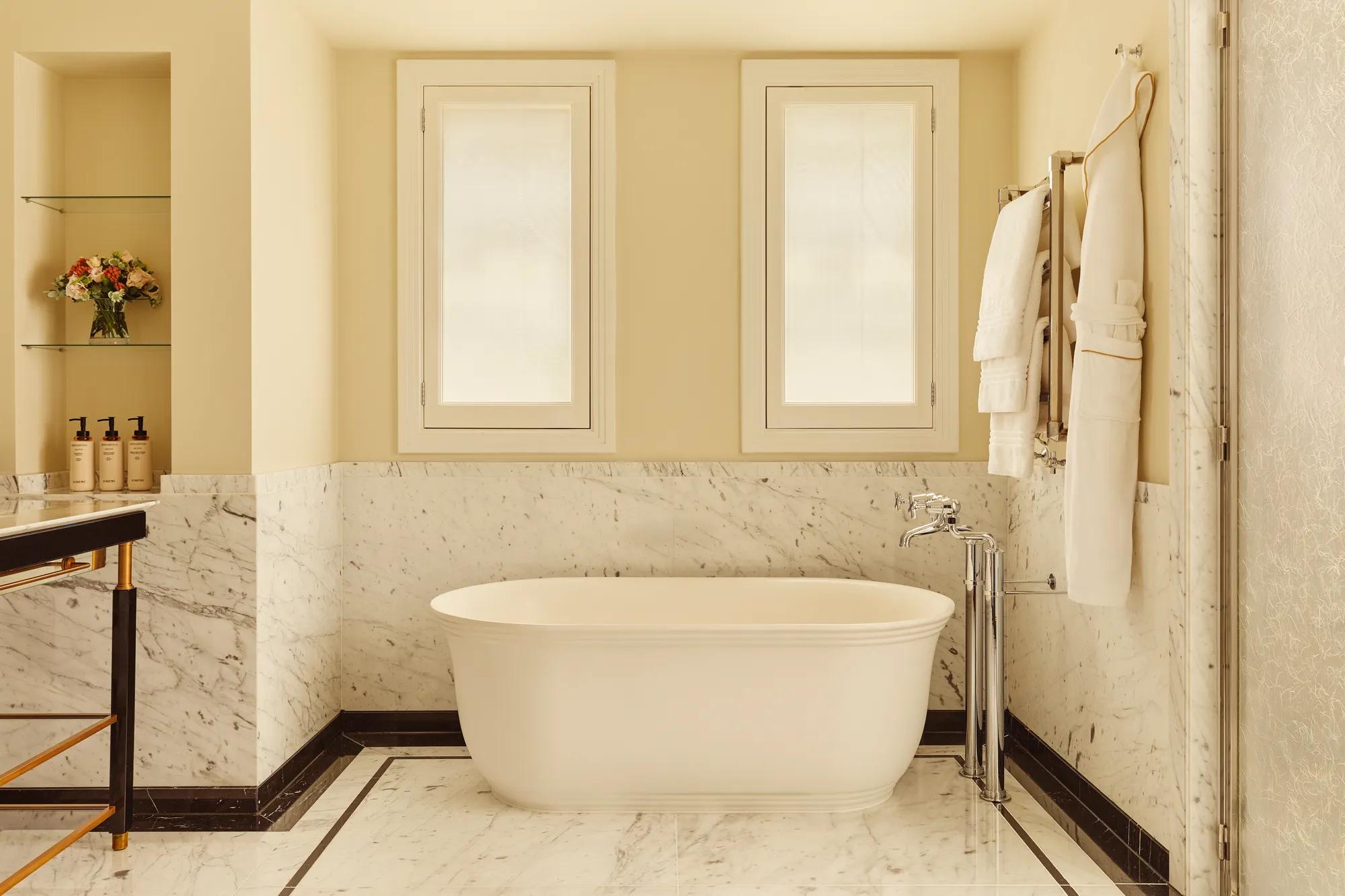 Bright marble bathroom with a freestanding white bathtub, chrome fixtures, and two frosted windows above, with towels and robes hanging neatly on the wall.