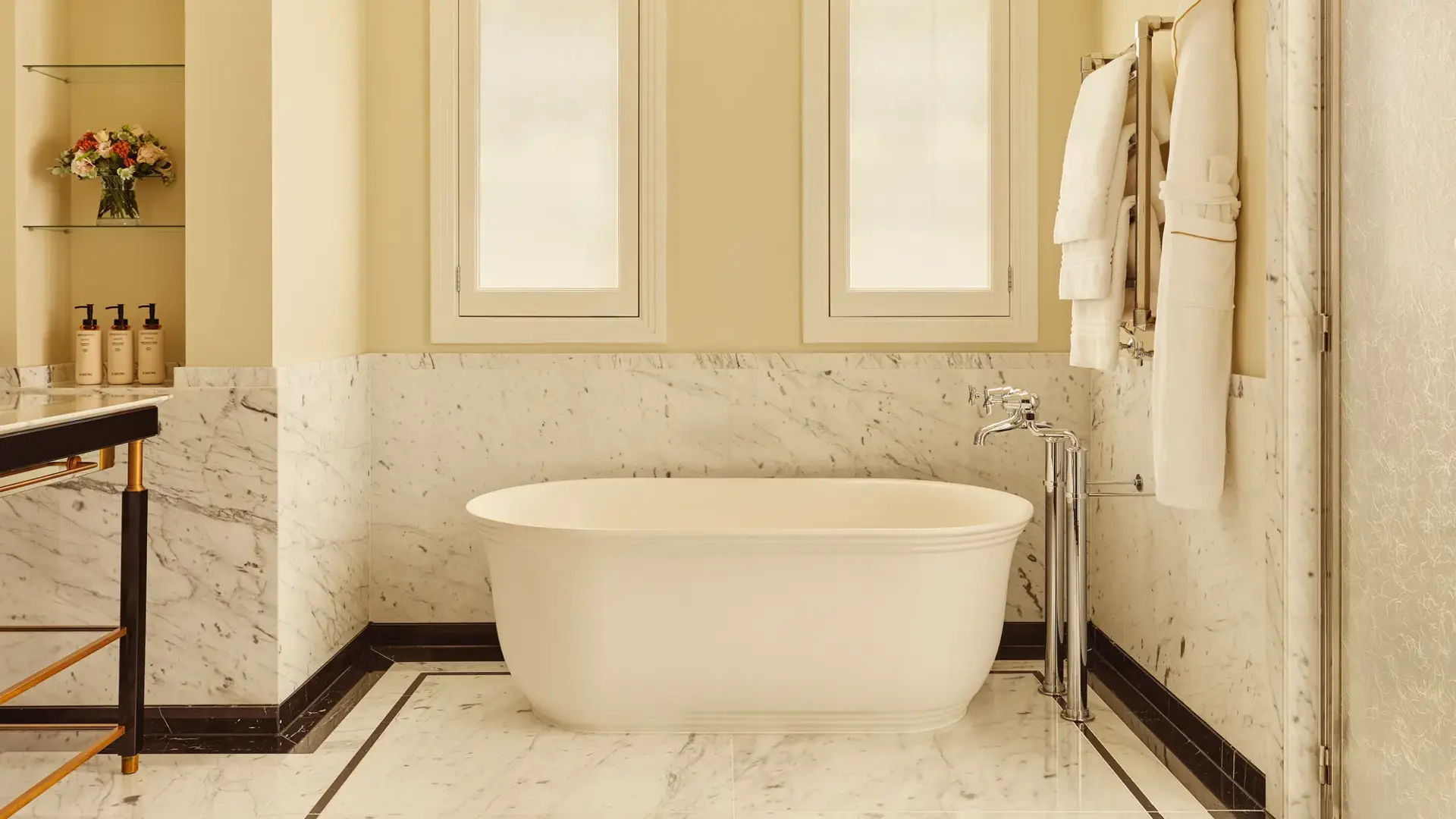 Bright marble bathroom with a freestanding white bathtub, chrome fixtures, and two frosted windows above, with towels and robes hanging neatly on the wall.