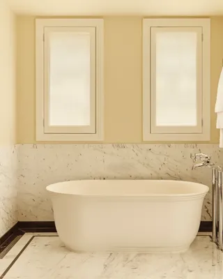 Bright marble bathroom with a freestanding white bathtub, chrome fixtures, and two frosted windows above, with towels and robes hanging neatly on the wall.