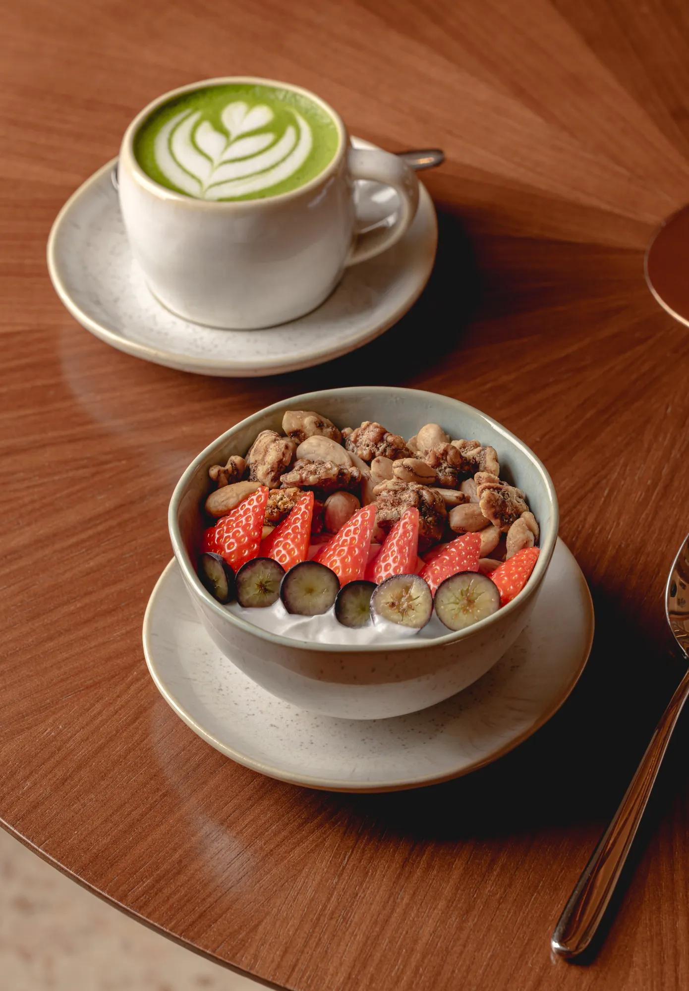 Bowl of yoghurt topped with strawberries, blueberries and granola, served with a matcha latte on a wooden table.