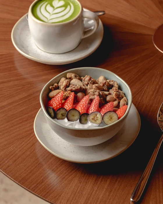 Bowl of yoghurt topped with strawberries, blueberries and granola, served with a matcha latte on a wooden table.