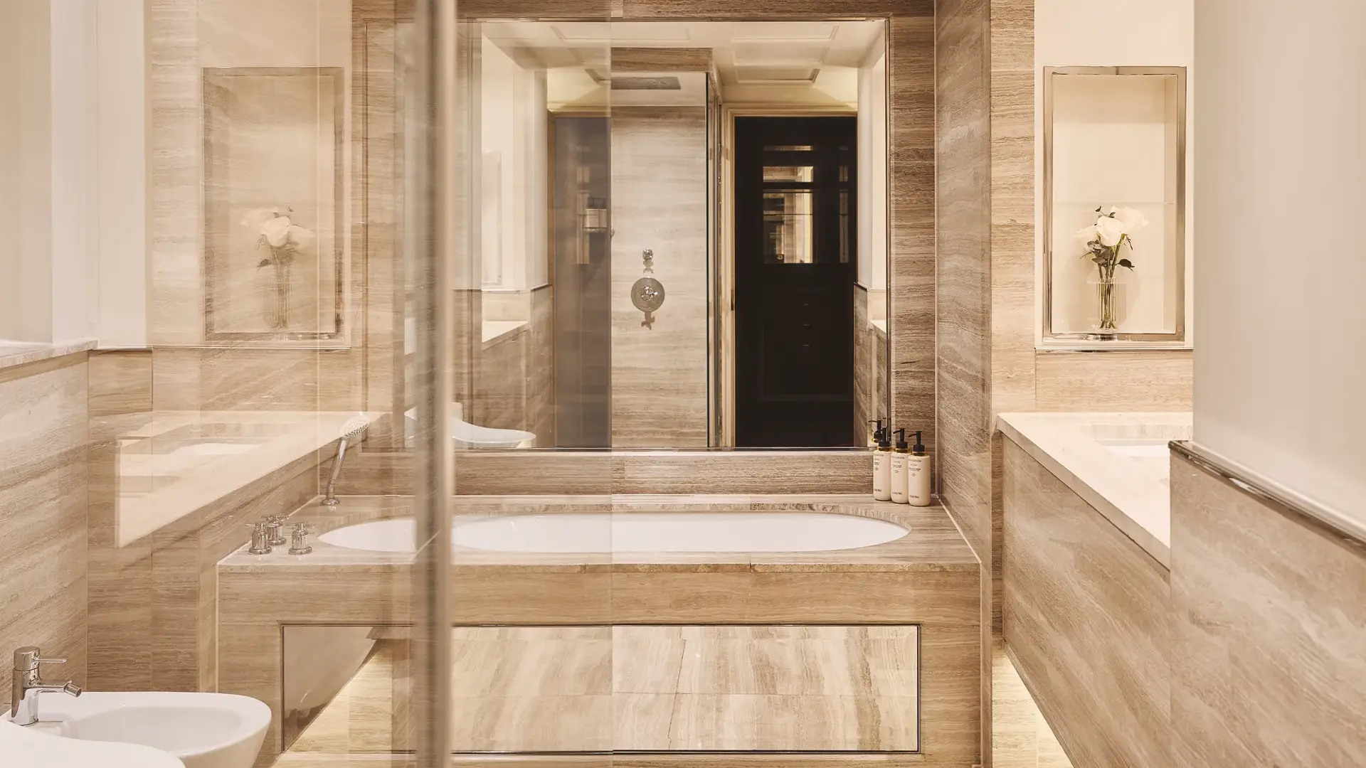 Beige marble bathroom with central bathtub, double vanity, glass shower, and bidet, softly lit with recessed lighting.