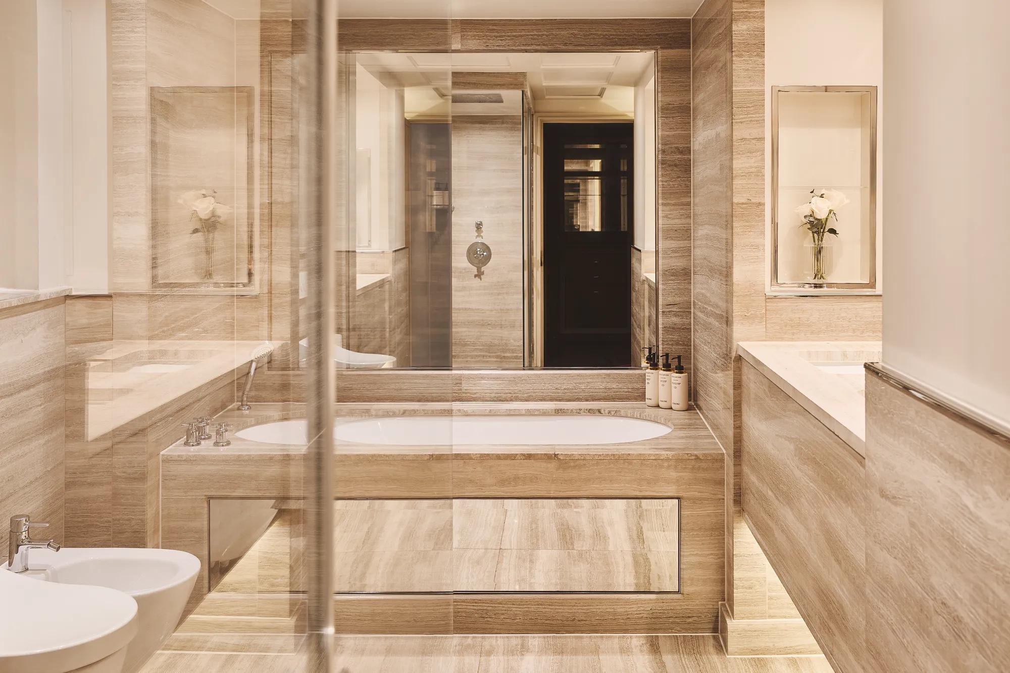 Beige marble bathroom with central bathtub, double vanity, glass shower, and bidet, softly lit with recessed lighting.