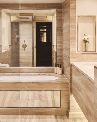 Beige marble bathroom with central bathtub, double vanity, glass shower, and bidet, softly lit with recessed lighting.