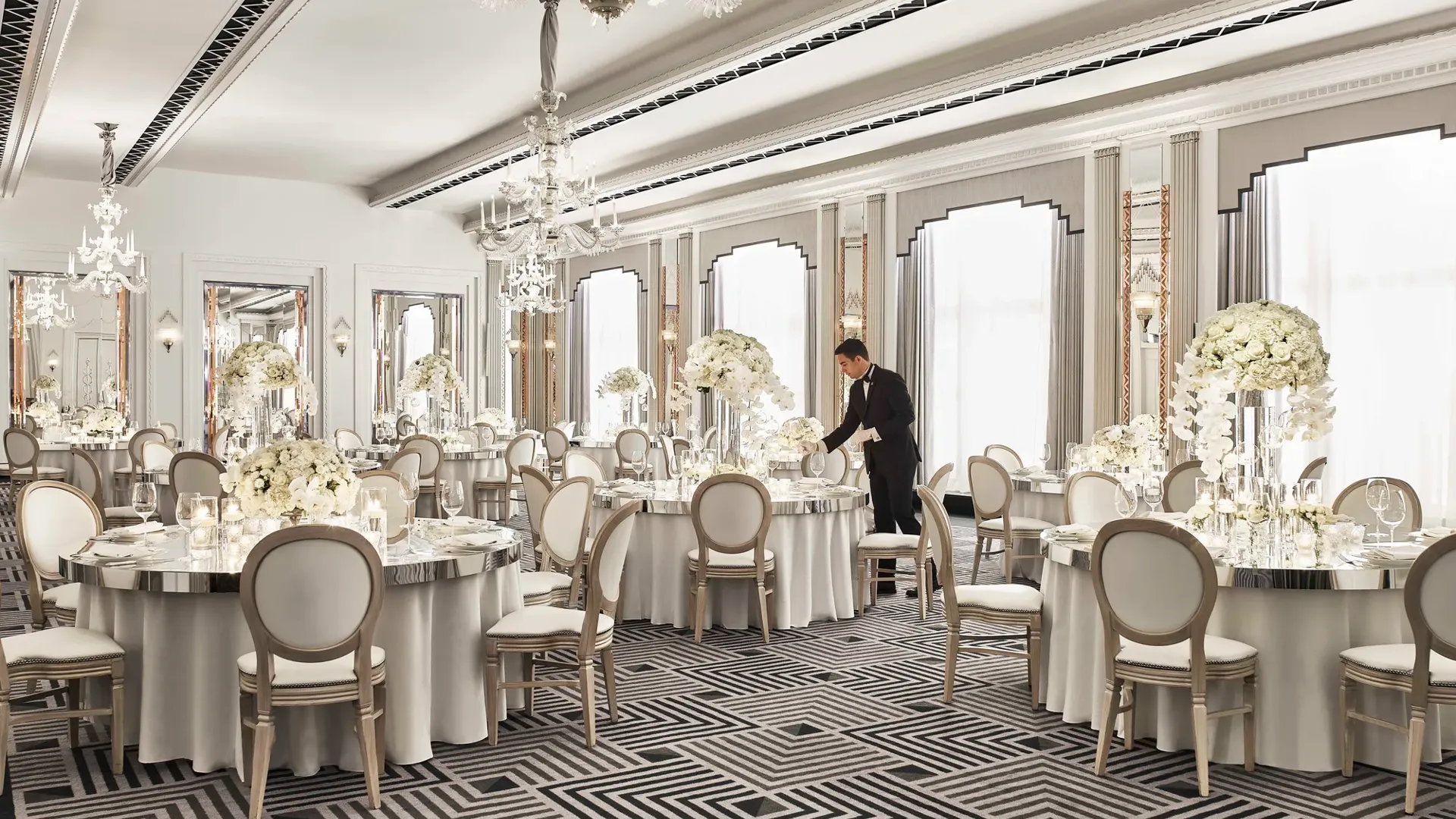 Elegant ballroom at Claridge’s set for a wedding reception, featuring round tables with white floral centrepieces, chandeliers, and mirrored accents in a refined Art Deco design.