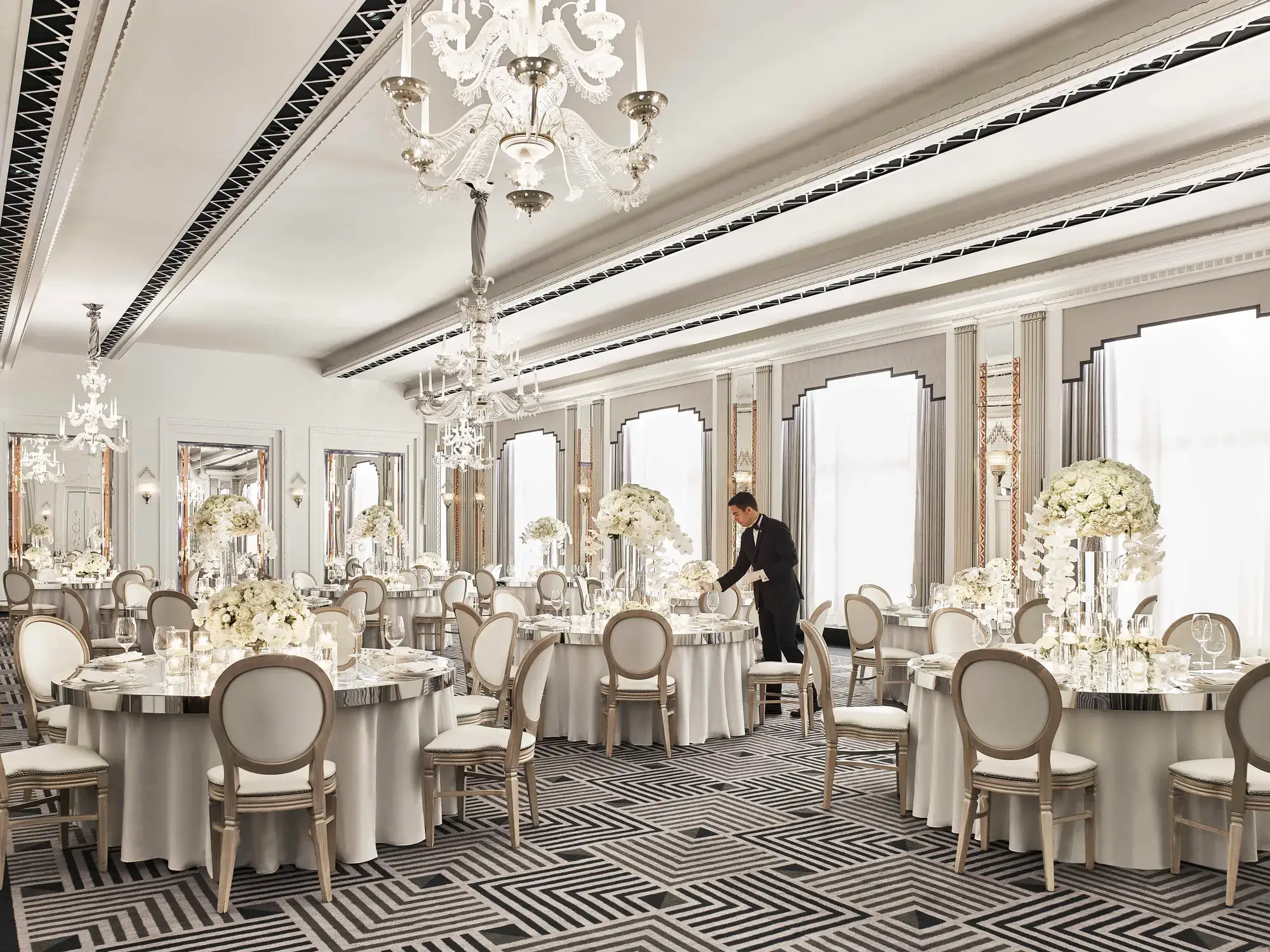 Elegant ballroom at Claridge’s set for a wedding reception, featuring round tables with white floral centrepieces, chandeliers, and mirrored accents in a refined Art Deco design.