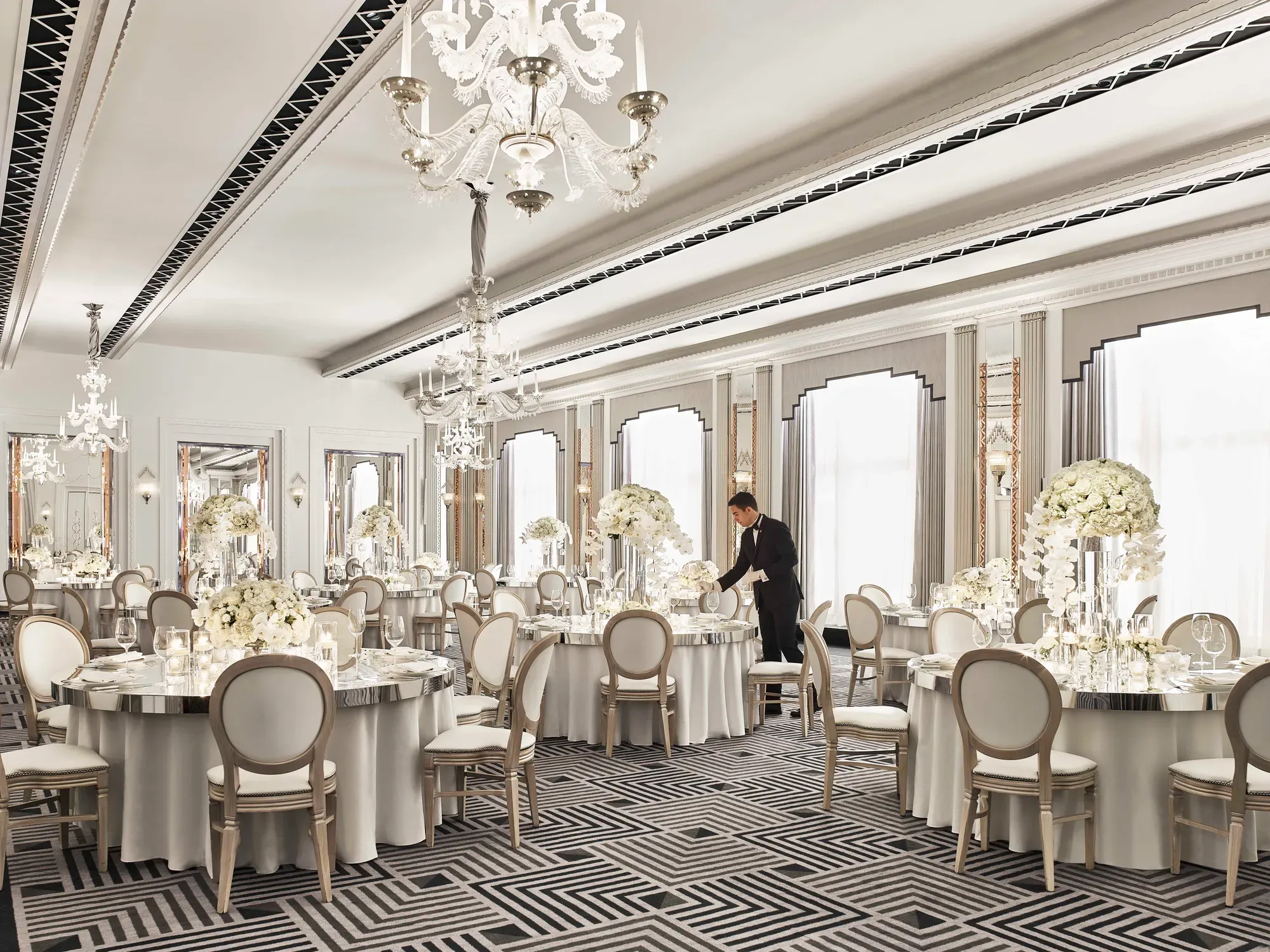 Elegant ballroom at Claridge’s set for a wedding reception, featuring round tables with white floral centrepieces, chandeliers, and mirrored accents in a refined Art Deco design.