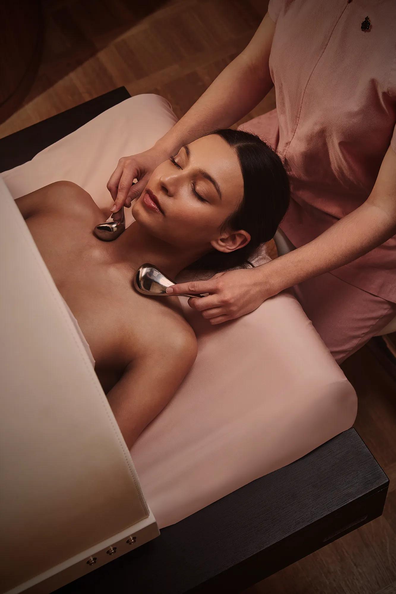 A woman receives a relaxing facial massage with cooling metal tools from a therapist in a softly lit treatment room at The Maybourne Riviera.