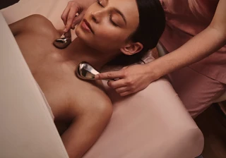 A woman receives a relaxing facial massage with cooling metal tools from a therapist in a softly lit treatment room at The Maybourne Riviera.