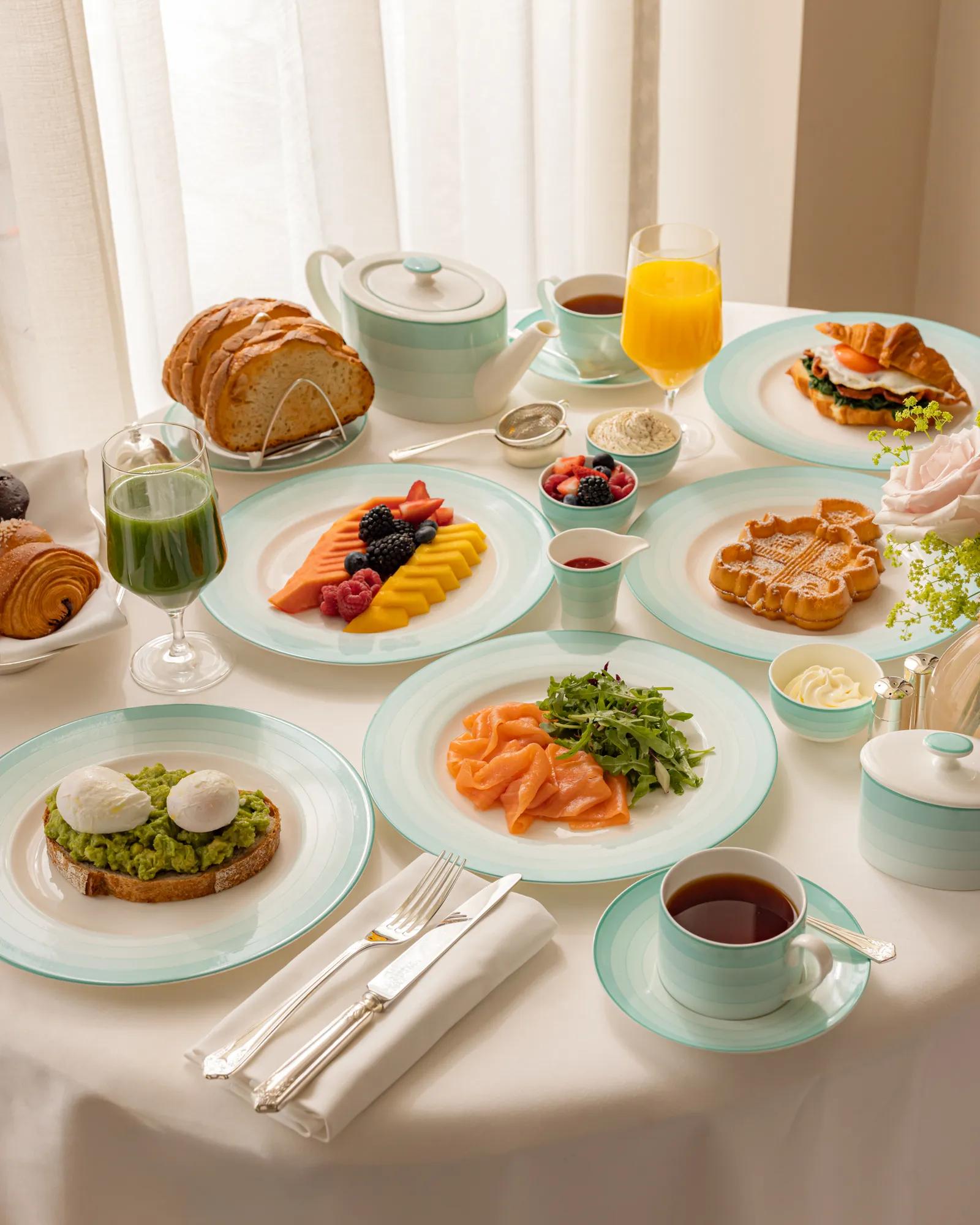 Elegant breakfast table set with smoked salmon, avocado toast with poached eggs, waffles, sliced fruit, pastries, and fresh juice. The tableware features mint-green and white china, arranged beside a teapot, coffee cup, and cutlery on a crisp white tablecloth.