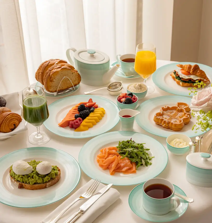 Elegant breakfast table set with smoked salmon, avocado toast with poached eggs, waffles, sliced fruit, pastries, and fresh juice. The tableware features mint-green and white china, arranged beside a teapot, coffee cup, and cutlery on a crisp white tablecloth.