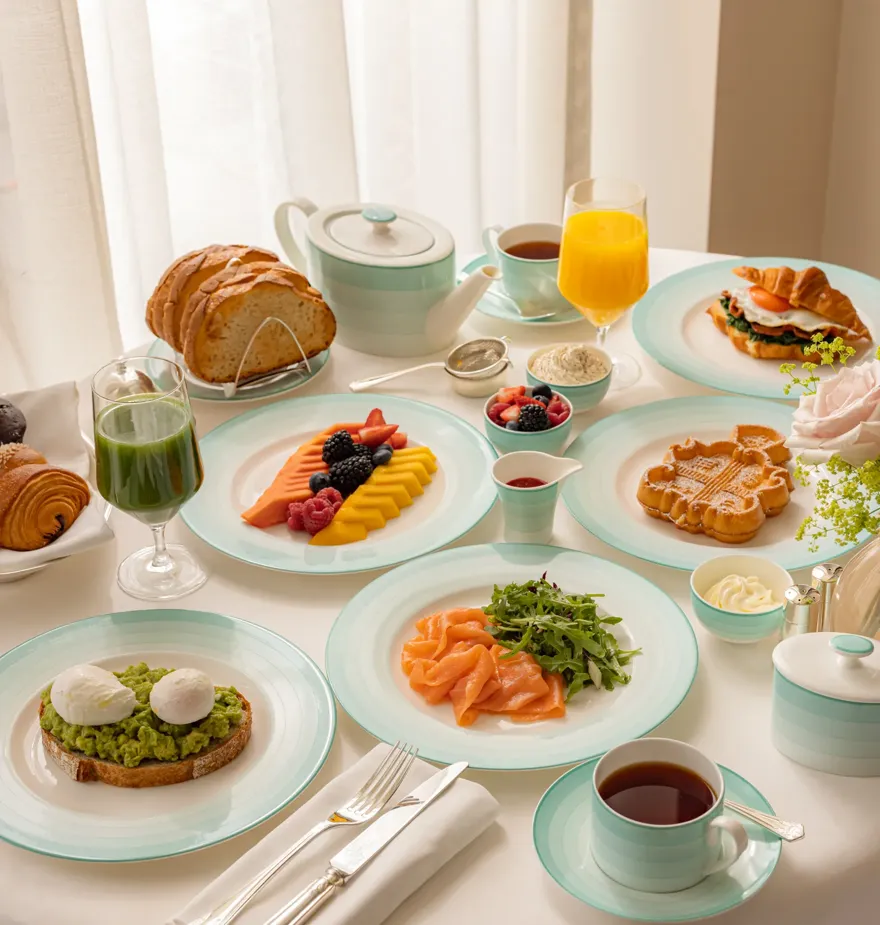 Elegant breakfast table set with smoked salmon, avocado toast with poached eggs, waffles, sliced fruit, pastries, and fresh juice. The tableware features mint-green and white china, arranged beside a teapot, coffee cup, and cutlery on a crisp white tablecloth.