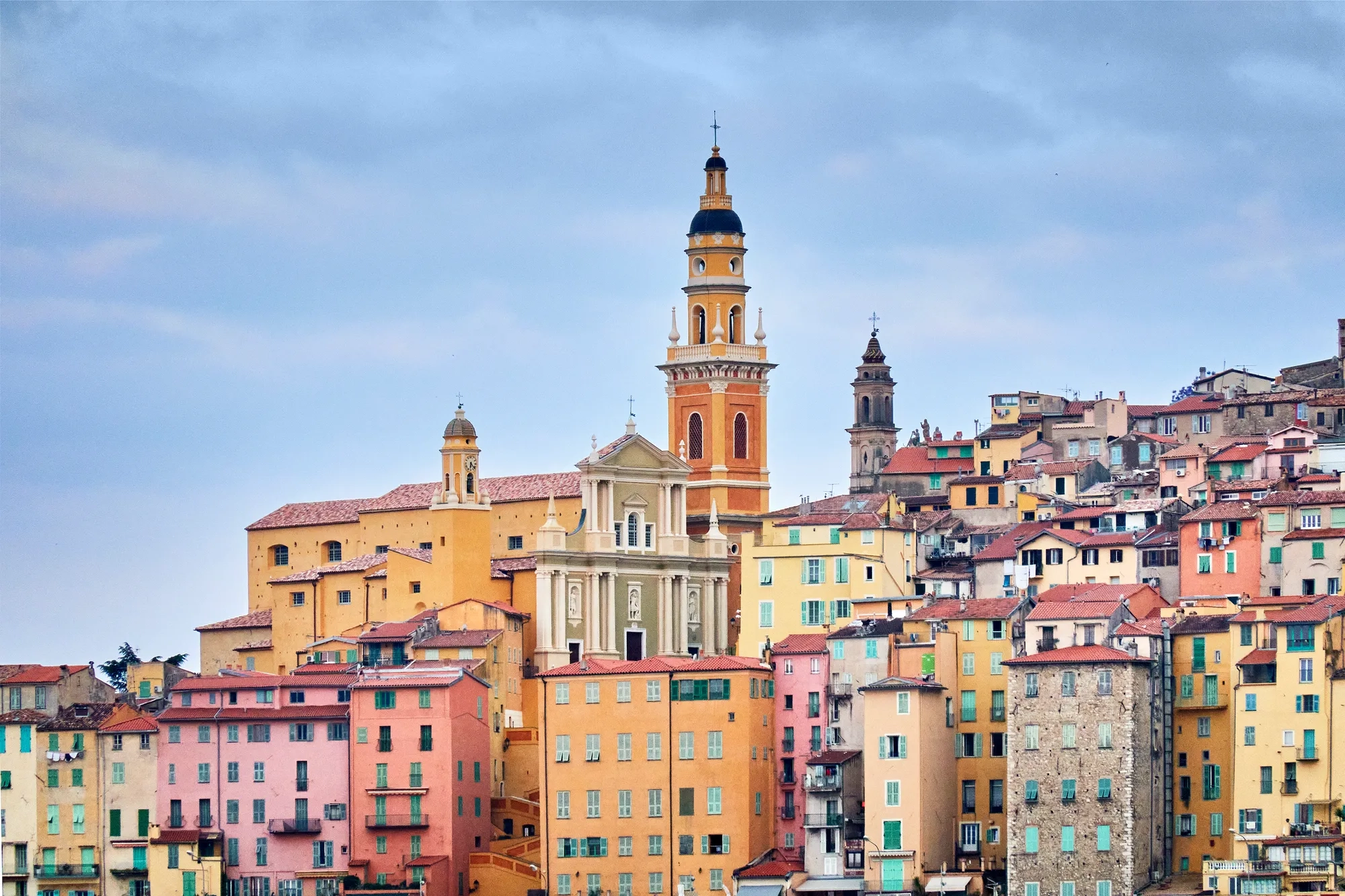 Colourful hillside town with pastel buildings and a prominent church tower rising above the rooftops under a cloudy sky.