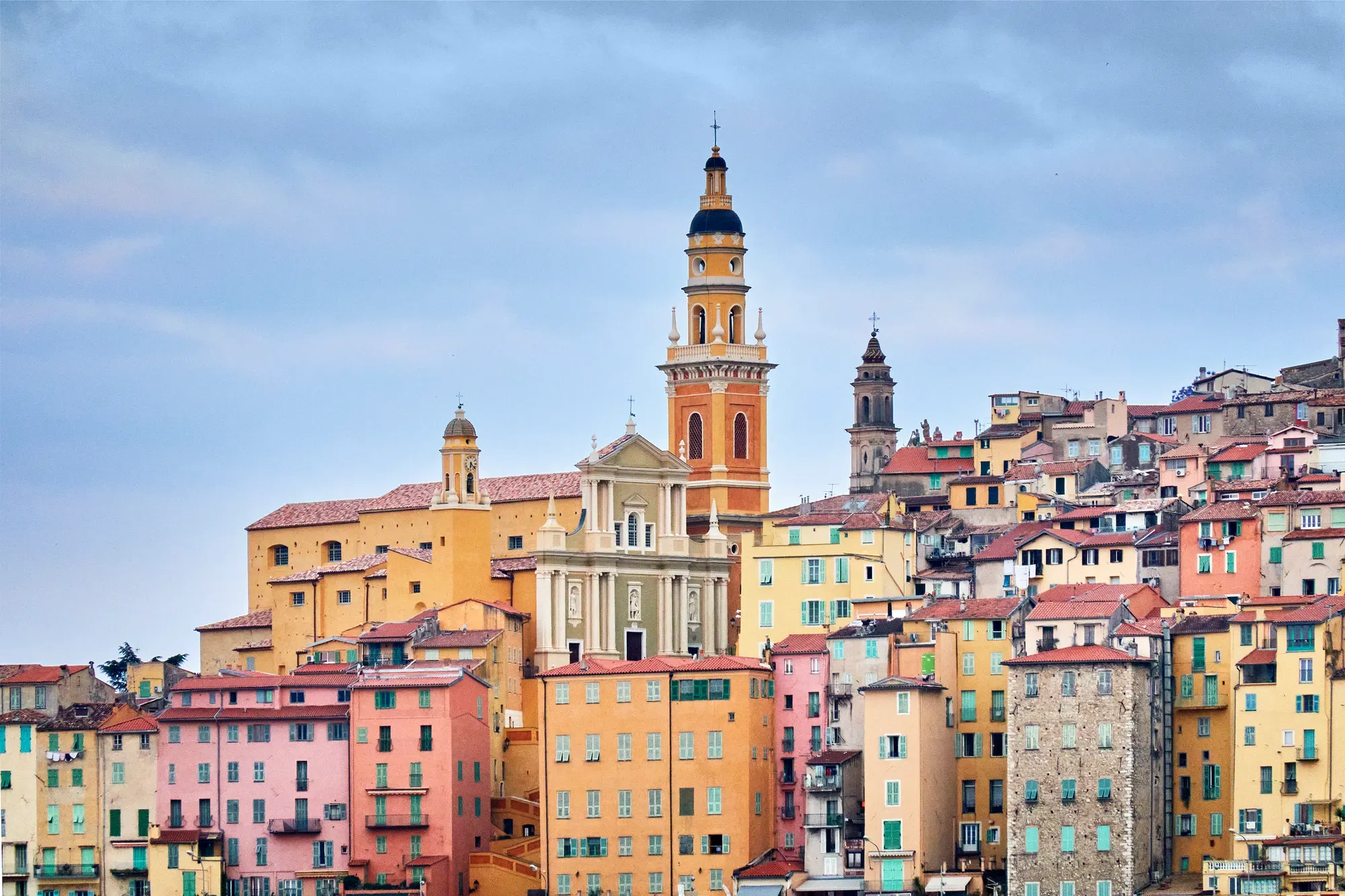 Colourful hillside town with pastel buildings and a prominent church tower rising above the rooftops under a cloudy sky.